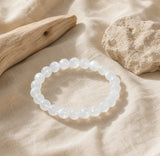 Bracelet en pierre de lune blanche sur du lin clair près de bois brut et d’une pierre mate.