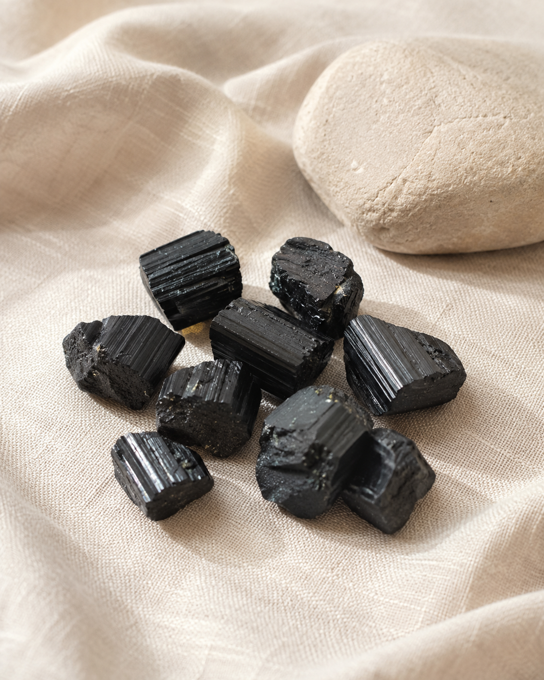 Ensemble de morceaux de tourmaline noire brute naturelle posés sur un tissu beige près d’un galet clair.